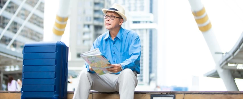 Senior man who purchased travel health insurance because he has existing medical conditions, with luggage sitting on staircase checking city map