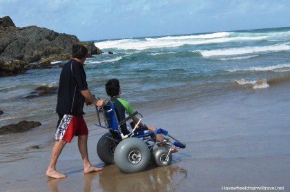 Photo of young man being pushed in wheelchair at the beach